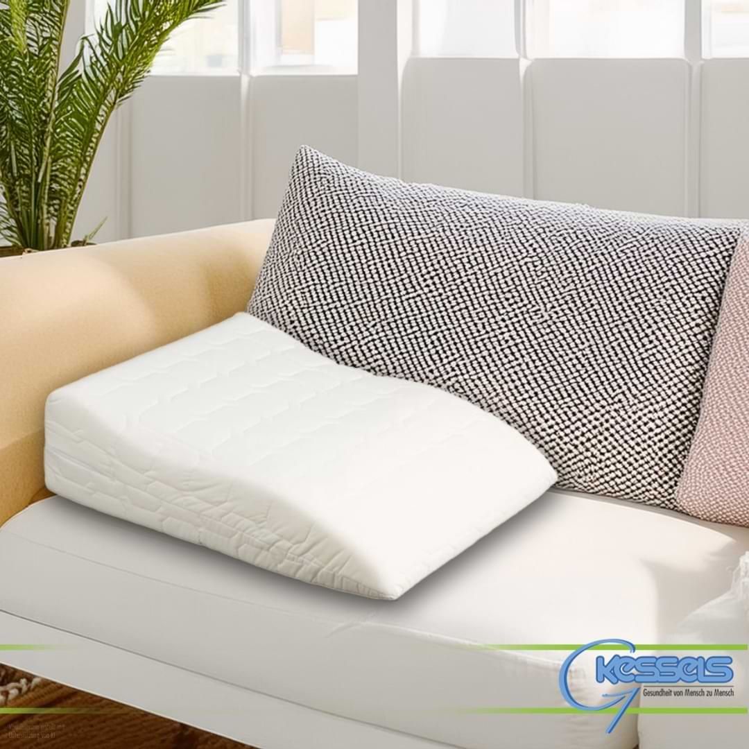 Ein weißes, ergonomisch geformtes Venenkissen zur Beinhochlagerung auf einem hellen Sofa.