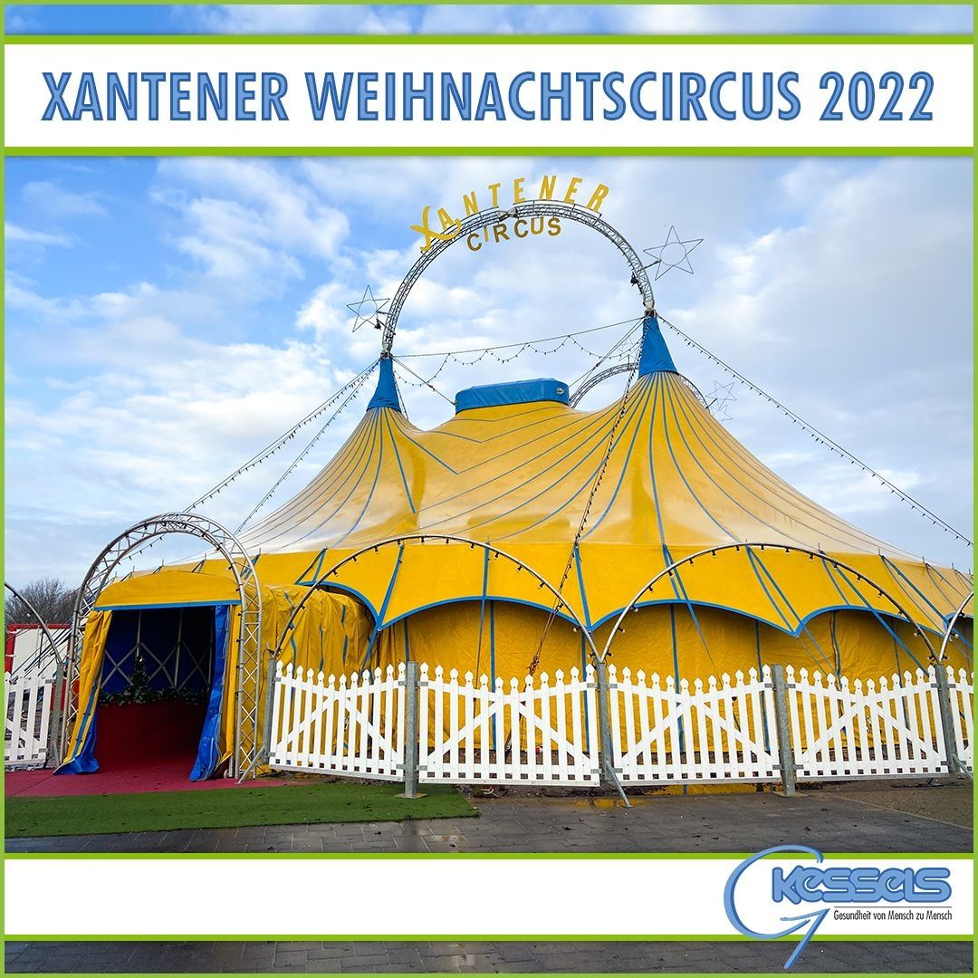 Leuchtend gelbes Zirkuszelt mit blauen Akzenten, umgeben von einem weißen Lattenzaun, unter einem strahlend blauen Himmel. Der Text oben lautet: Xantener Weihnachtscircus 2022.