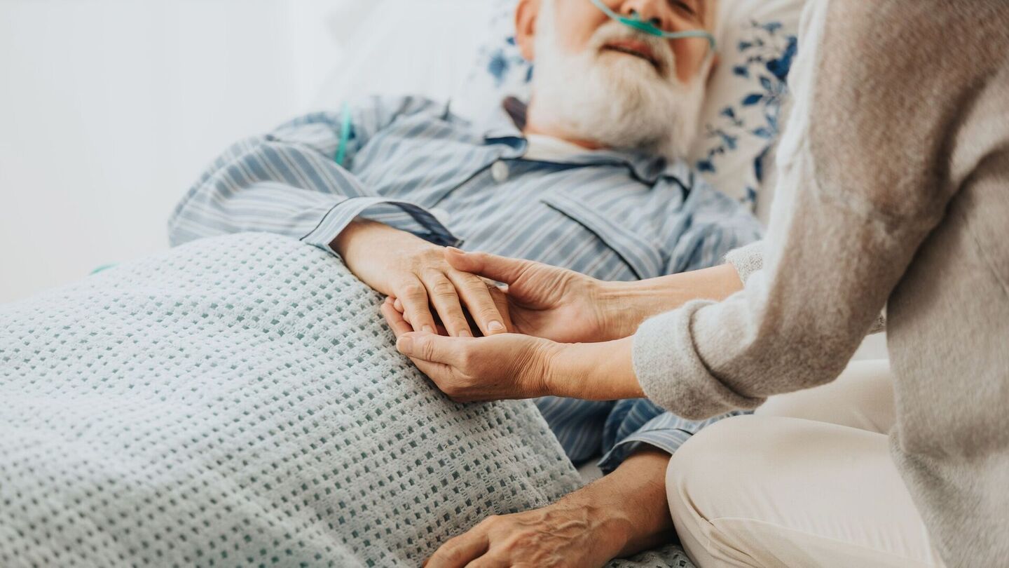 Eine Pflegekraft hält sanft die Hand eines Patienten, der im Bett ruht, umgeben von weicher Bettwäsche, und vermittelt ein Gefühl von Mitgefühl und Trost.