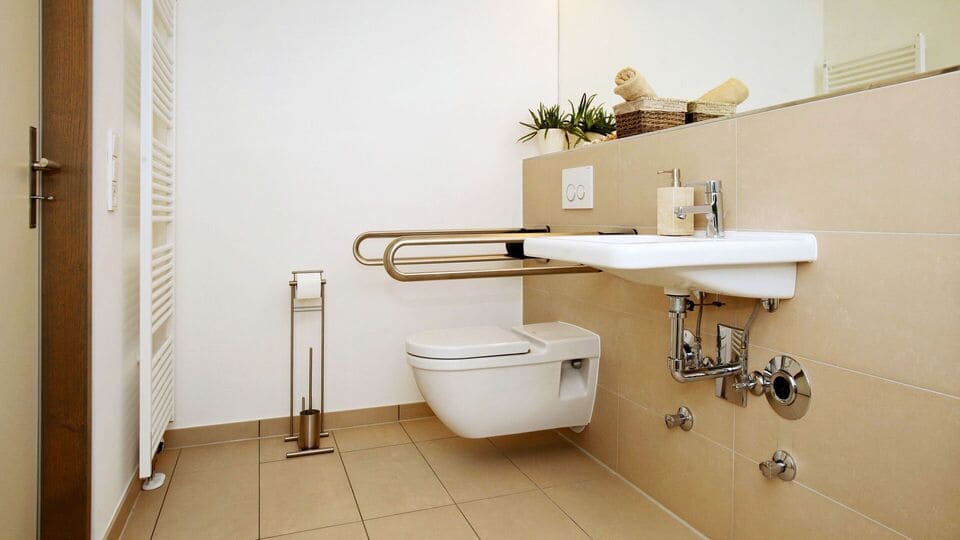Modernes, barrierefreies Badezimmer mit wandmontierter Toilette, Haltegriffen, Waschbecken und minimalistischer Einrichtung mit Pflanzen und Handtüchern.